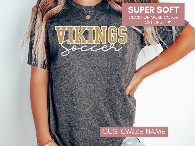 May include: A gray t-shirt with the text "VIKINGS Soccer" in gold and white lettering. The text "SUPER SOFT" and "CLICK FOR MORE COLOR OPTIONS" is displayed in pink at the top of the image. The text "CUSTOMIZE NAME" is displayed in pink at the bottom of the image.
