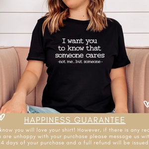 以下が含まれることがあります： 「I want you to know that someone cares -not me, but someone-」という白文字がプリントされた黒いTシャツ。Tシャツは人が着ています。画像の下部には「HAPPINESS GUARANTEE」という文字があります。