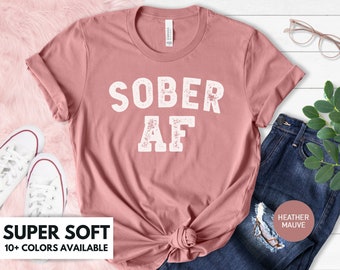 Puede incluir: Camiseta color malva con la frase "SOBER AF" en letras blancas desgastadas. La camiseta está anudada a la cintura y combinada con vaqueros azules. Las palabras "SUPER SOFT" y "10+ COLORS AVAILABLE" se muestran en un cuadro blanco.