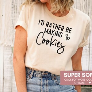 May include: A beige t-shirt with the text "I'd rather be making cookies" in black lettering. The shirt is super soft and available in multiple colors.
