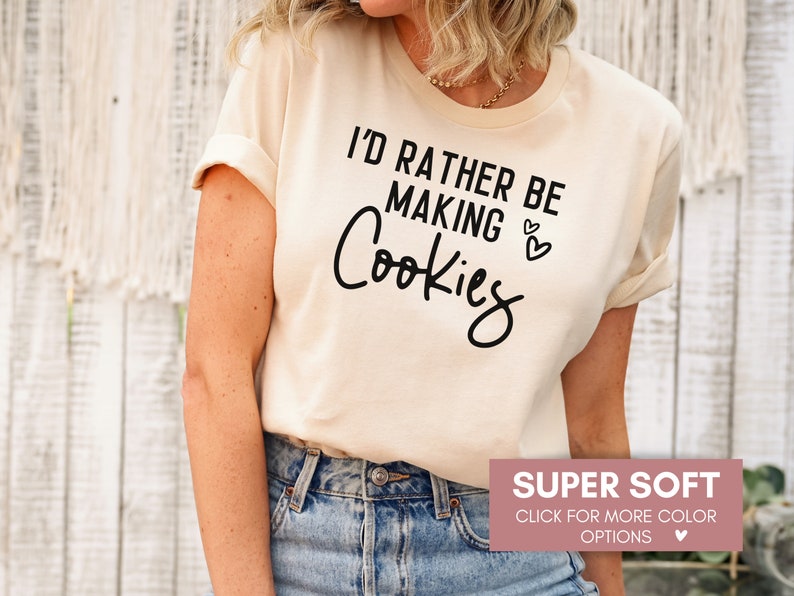 May include: A beige t-shirt with the text "I'd rather be making cookies" in black lettering. The shirt is super soft and available in multiple colors.