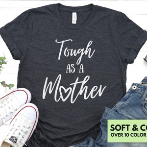 K&ouml;nnte beinhalten: Ein dunkelgraues T-Shirt mit dem Text "Tough as a Mother" in wei&szlig;en Buchstaben mit einem kleinen Herzen in der Mitte des Wortes "Mother". Das Shirt ist weich und bequem und in &uuml;ber 10 Farben erh&auml;ltlich.