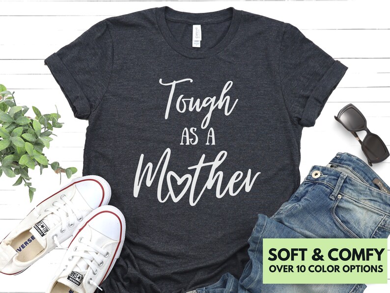 K&ouml;nnte beinhalten: Ein dunkelgraues T-Shirt mit dem Text "Tough as a Mother" in wei&szlig;en Buchstaben mit einem kleinen Herzen in der Mitte des Wortes "Mother". Das Shirt ist weich und bequem und in &uuml;ber 10 Farben erh&auml;ltlich.