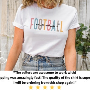 Op de afbeelding: Wit T-shirt met de tekst "FOOTBALL mama" in kleurrijke letters met een klein hartje aan het einde. Het shirt wordt gedragen door een persoon met een blauwe jeans.