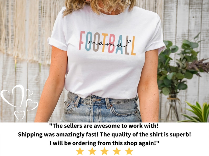 Op de afbeelding: Wit T-shirt met de tekst "FOOTBALL mama" in kleurrijke letters met een klein hartje aan het einde. Het shirt wordt gedragen door een persoon met een blauwe jeans.