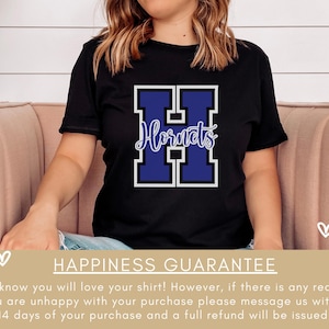 Puede incluir: Camiseta negra con una gran letra "H" azul y blanca con la palabra "Hornets" escrita en blanco dentro de la letra. La camiseta la lleva puesta una persona sentada en un sof&aacute; rosa. El texto "Happiness Guarantee" est&aacute; en la parte inferior de la imagen.