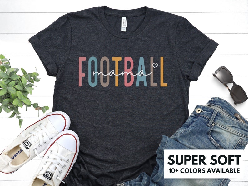 Op de afbeelding: Een donkergrijs t-shirt met de tekst "FOOTBALL mama" in regenboogkleuren met een klein hartje na het woord "mama".