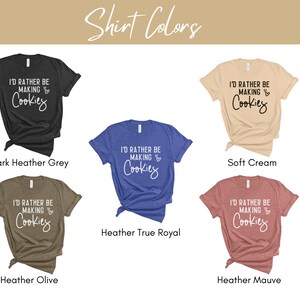 May include: Five t-shirts in different colors with the text "I'd rather be making cookies" printed on the front. The colors are dark heather grey, soft cream, heather true royal, heather olive, and heather mauve.