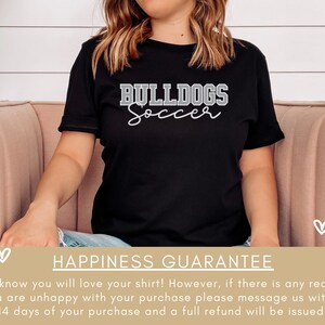 May include: Black t-shirt with white text that reads "BULLDOGS Soccer". The shirt is worn by a person sitting on a pink couch. The background is a white wood panel wall.