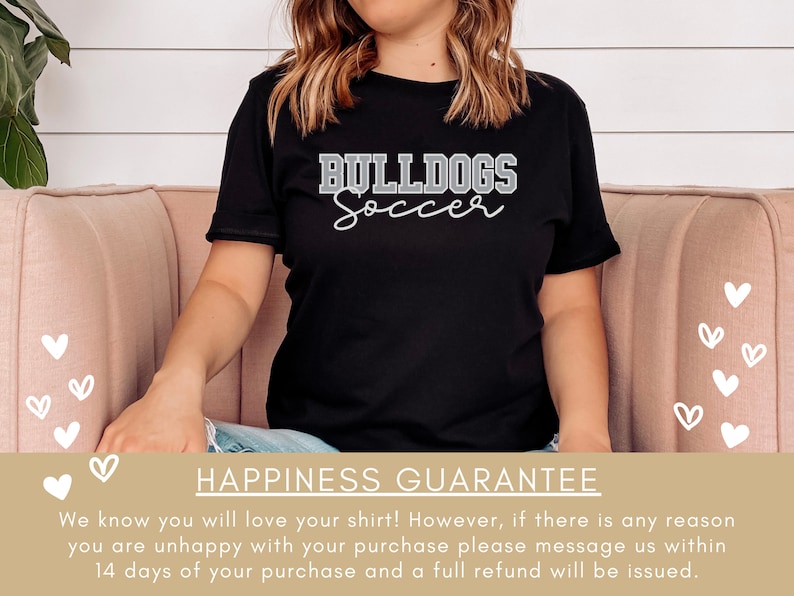 May include: Black t-shirt with white text that reads "BULLDOGS Soccer". The shirt is worn by a person sitting on a pink couch. The background is a white wood panel wall.