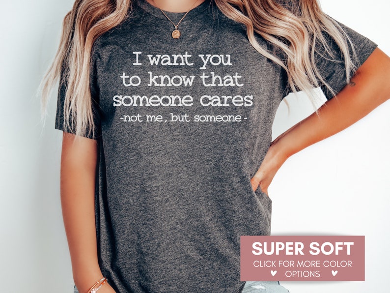 以下が含まれることがあります： 「I want you to know that someone cares - not me, but someone -」という白文字がプリントされた濃いグレーのTシャツ。下部には「SUPER SOFT」と「CLICK FOR MORE COLOR OPTIONS」と書かれたピンク色のボックスがあります。