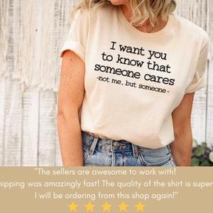 以下が含まれることがあります： 「I want you to know that someone cares - not me, but someone -」というテキストがプリントされたクリーム色のTシャツ。袖はロールアップされ、カジュアルなフィット感。テキストは黒色です。
