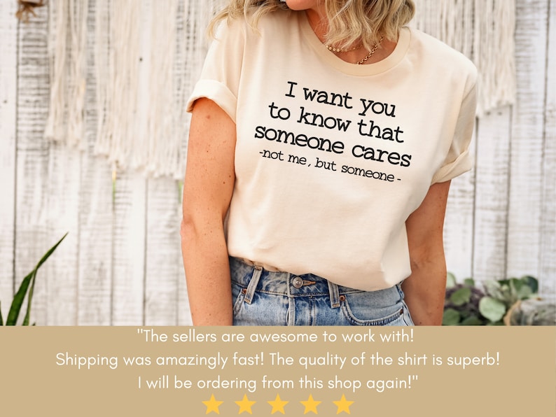 以下が含まれることがあります： 「I want you to know that someone cares - not me, but someone -」というテキストがプリントされたクリーム色のTシャツ。袖はロールアップされ、カジュアルなフィット感。テキストは黒色です。