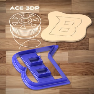 Puede incluir: Un cortador de galletas de plástico azul en forma de la letra B. El cortador tiene tres pequeños recortes rectangulares en el interior. El cortador está sobre una superficie de madera.