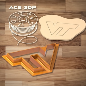 May include: A gold-colored plastic cookie cutter in the shape of the letters "VT". The cutter is on a wooden surface. There is a white outline of the same design on a beige surface and a spool of white filament.