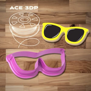 Puede incluir: Dos cortadores de galletas, uno con forma de gafas de sol y el otro con forma de gafas de buceo. El cortador de gafas de sol es amarillo y el cortador de gafas de buceo es rosa.