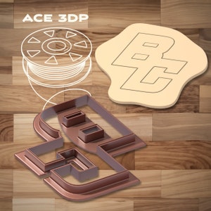 Puede incluir: Un cortador de galletas de plástico marrón en forma de la letra "B" con una "C" más pequeña en su interior. El cortador de galletas está sobre una superficie de madera. El texto "ACE 3DP" es visible en la esquina superior izquierda de la imagen. También se puede ver un contorno blanco de una bobina de filamento en la esquina superior izquierda de la imagen.