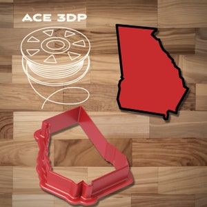 May include: Red plastic cookie cutter in the shape of the state of Georgia. The cookie cutter is on a wooden surface. The image also shows a spool of white filament and a black outline of the state of Georgia.