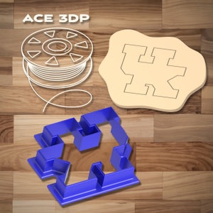 Puede incluir: Un cortador de galletas de plástico azul en forma de la letra "K" con un diseño decorativo. El cortador de galletas está sobre una superficie de madera. La imagen también muestra una bobina de filamento blanco y un cortador de galletas de madera en forma de la letra "K".