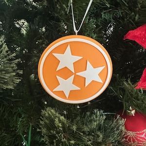 Puede incluir: Adorno navide&ntilde;o naranja con un contorno blanco y tres estrellas blancas en forma de la bandera del estado de Tennessee.