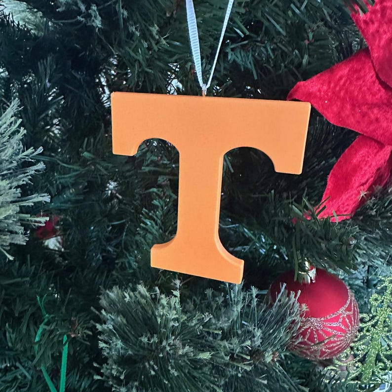 Puede incluir: Adorno de Navidad de la letra T naranja colgado de un &aacute;rbol de Navidad verde.