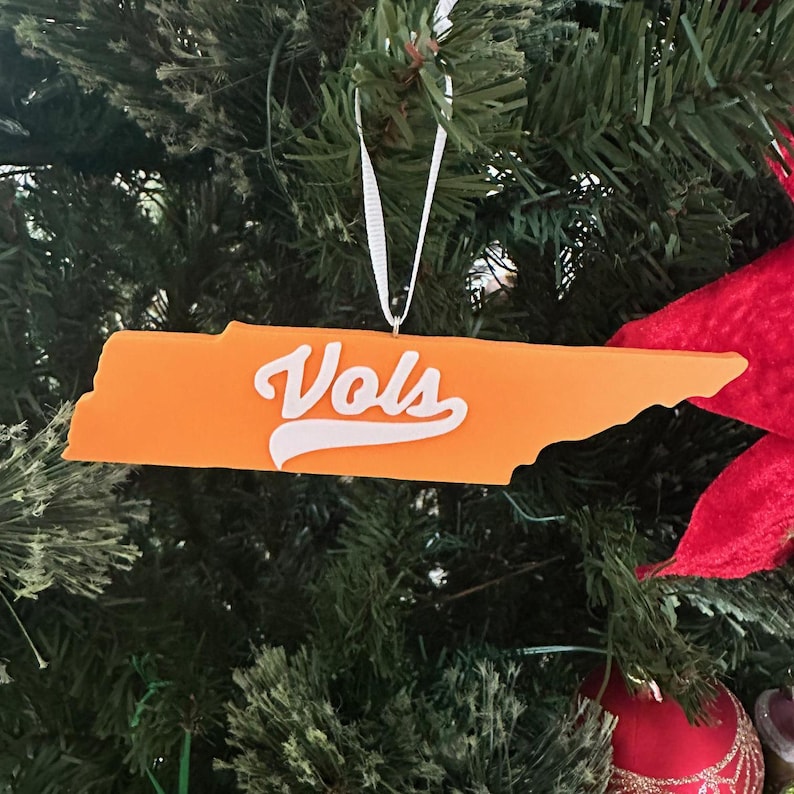 Puede incluir: Adorno de Navidad de color naranja en forma del estado de Tennessee con la palabra "Vols" en letras blancas.