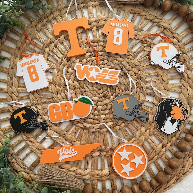 Puede incluir: Una colecci&oacute;n de adornos navide&ntilde;os de color naranja y blanco con varios logotipos y dise&ntilde;os de los Volunteers de la Universidad de Tennessee, incluyendo la mascota del equipo, un casco de f&uacute;tbol americano, una camiseta y el estado de Tennessee. Los adornos se muestran en una estera de paja tejida.