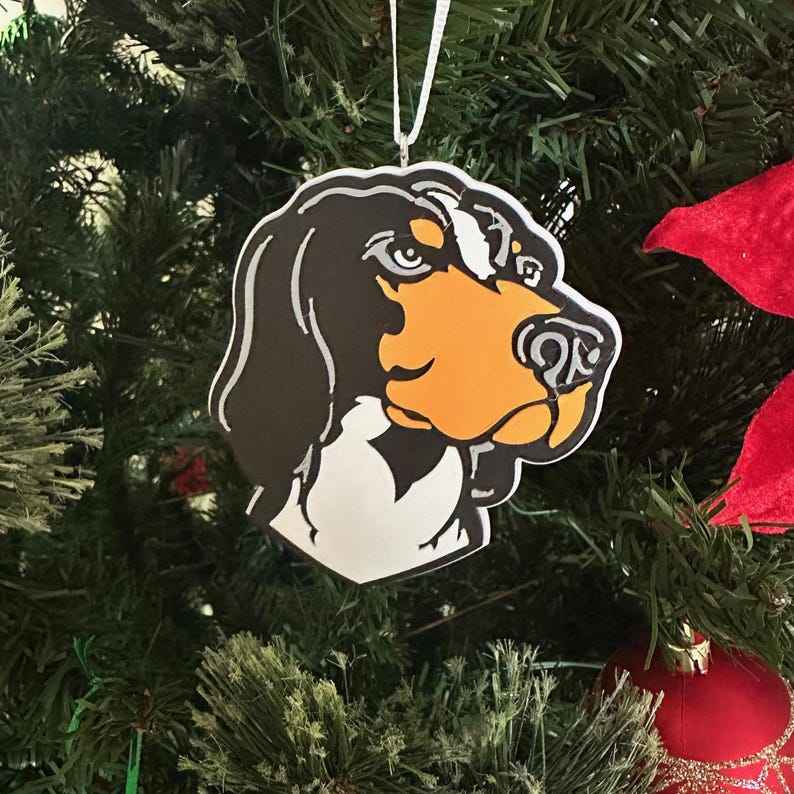Puede incluir: Un adorno navide&ntilde;o de color blanco, negro y naranja con forma de cabeza de perro. El adorno est&aacute; colgado de una cuerda blanca en un &aacute;rbol de Navidad.