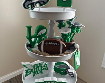Puede incluir: Una bandeja blanca de tres niveles decorada con artículos temáticos de los Philadelphia Eagles. Incluye un balón de fútbol, una camiseta que dice "HURTS 1", el logotipo del equipo y las frases "FLY EAGLES FLY" y "GO BIRDS".