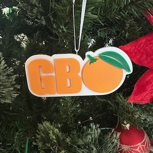 Puede incluir: Adorno navide&ntilde;o blanco con las letras "GBO" en naranja y una naranja estilizada con una hoja verde en el lado derecho.
