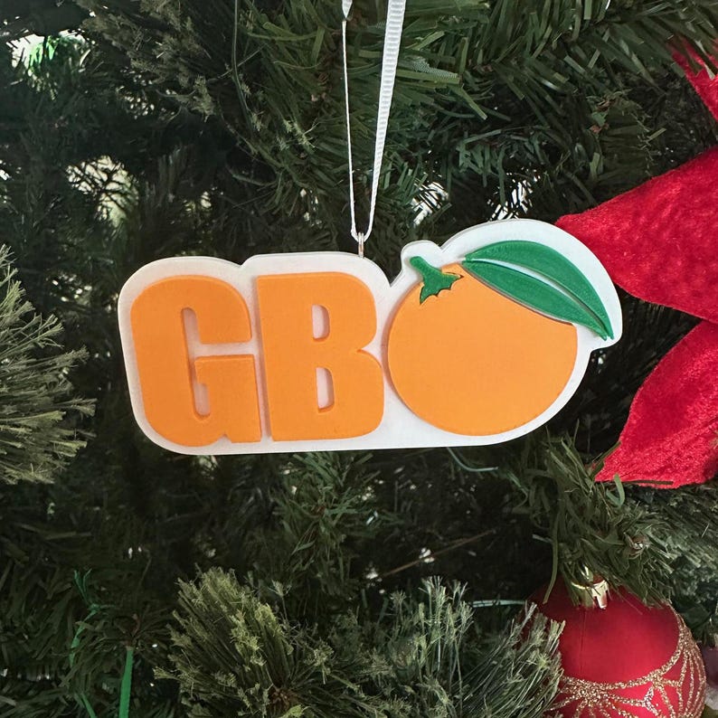 Puede incluir: Adorno navide&ntilde;o blanco con las letras "GBO" en naranja y una naranja estilizada con una hoja verde en el lado derecho.