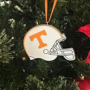 Puede incluir: Adorno navide&ntilde;o blanco y naranja con forma de casco de f&uacute;tbol americano con una gran "T" naranja en la parte delantera.