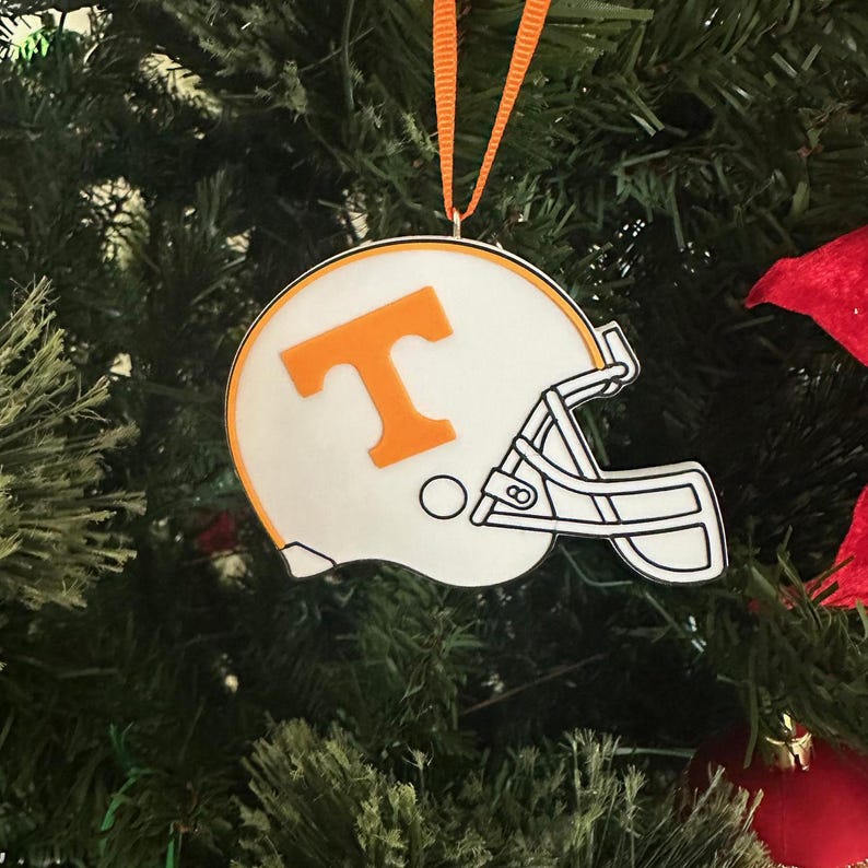 Puede incluir: Adorno navide&ntilde;o blanco y naranja con forma de casco de f&uacute;tbol americano con una gran "T" naranja en la parte delantera.