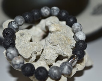 May include: A beaded bracelet featuring a mix of black lava rock and gray patterned stone beads. The bracelet is strung on a stretch cord and has a silver-toned metal clasp. The bracelet is displayed on a piece of white coral.