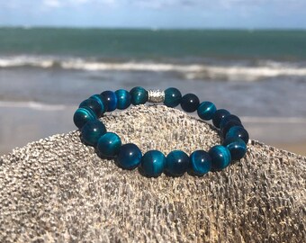 Puede incluir: Un brazalete de cuentas de ojo de tigre azul con un acento de cuentas de plata. El brazalete está sobre un trozo de madera a la deriva con un fondo borroso de una playa y el océano.
