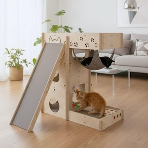 May include: A multi-level cat tree made of light-colored wood, featuring a gray slide, multiple openings, a hammock, and a scratching pad. Two black cats rest in the hammock, while an orange tabby plays with a toy. The cat tree is decorated with paw prints and cat silhouettes.