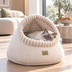 May include: A cream-coloured, dome-shaped pet bed made of ribbed corduroy fabric with a thick, rounded edge. A fluffy cat with blue eyes rests inside the cosy, enclosed space. A small rectangular brown label on the front features the text PRASKA.