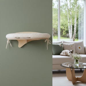May include: A wall-mounted cat shelf with a cream-colored, ribbed cushion. The shelf is made of light-colored wood and has a curved design. Cream-colored ties are attached to the bottom of the shelf. The shelf is mounted on a green wall.