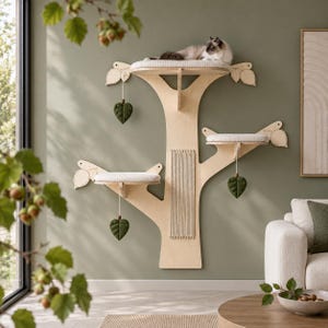 May include: A wall-mounted cat tree made of light-colored wood, featuring three tiered platforms with white cushions, a central vertical scratching post, and three hanging green leaf-shaped toys. A cat rests on the top platform.