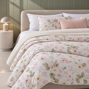 Pode incluir: Um conjunto de cama composto por um edredão branco e fronhas a condizer com uma estampa floral suave em tons de rosa e verde sálvia. O conjunto inclui uma almofada retangular cor-de-rosa claro lisa e um acabamento em lã sherpa creme.