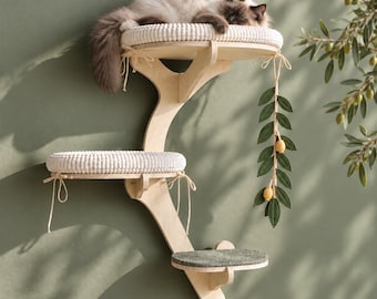 May include: A wall-mounted cat tree featuring a light wood frame with three tiered platforms. Two upper platforms have white textured cushions tied with strings, while the lowest platform has a dark gray mat. A decorative felt olive branch hangs from the top.
