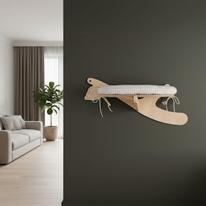 May include: A wooden cat shelf with a white, woven bed. The shelf is mounted on a dark green wall. The shelf has a unique airplane-like design with a curved shape and is secured with white ties.