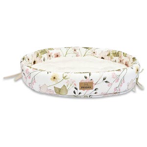 Può includere: Lettino ovale per animali domestici con un motivo floreale bianco con fiori rosa e gialli e foglie verdi. Il letto ha un interno bianco morbido e una piccola etichetta marrone con la scritta "PRASKA". I lacci beige sono attaccati ai lati.