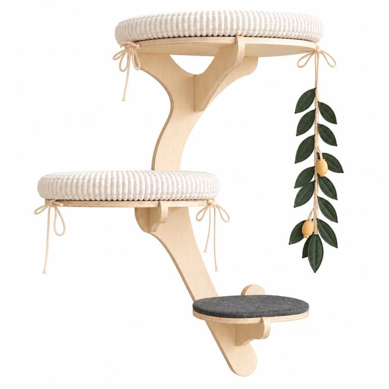 May include: A wall-mounted cat tree featuring a light wood frame with three tiered platforms. The top two platforms have round, textured white cushions with tie-fasteners. The lowest platform has a dark gray cushion. A hanging green leaf toy with yellow accents is attached.