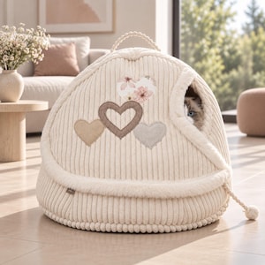 Pode incluir: Uma cama para animais de estimação em veludo cotelê cor de creme, em forma de cúpula e com rebordo em pelo sintético. O exterior apresenta três aplicações em forma de coração, uma com padrão floral, e uma pega. Um gato espreita pela abertura.
