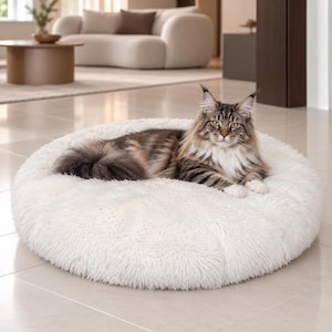 May include: A fluffy, round, off-white pet bed with a thick, shaggy texture. A long-haired, brown and black tabby cat with prominent ear tufts is lying down in the center of the bed, facing forward with its paws tucked underneath its body.
