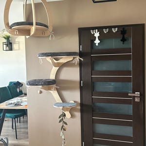 May include: A wall-mounted cat furniture system featuring a suspended ceiling perch and a multi-level climbing structure with three platforms. Each platform is fitted with a dark gray textured cushion, and a hanging leaf toy is attached.