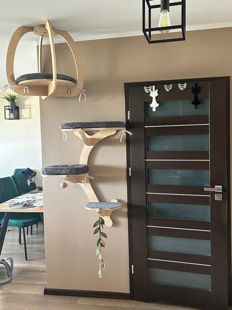 May include: A wall-mounted cat furniture system featuring a suspended ceiling perch and a multi-level climbing structure with three platforms. Each platform is fitted with a dark gray textured cushion, and a hanging leaf toy is attached.