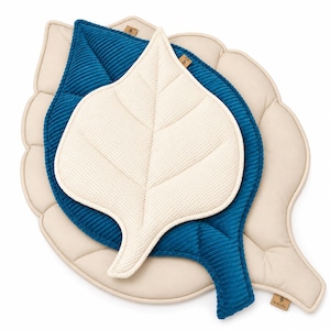 Puede incluir: Tres cojines con forma de hoja apilados. El cojín inferior es beige con superficie lisa, el central es de pana azul profundo y el superior es de tela color crema texturizada. Cada cojín presenta detalles de nervaduras cosidas y una etiqueta.