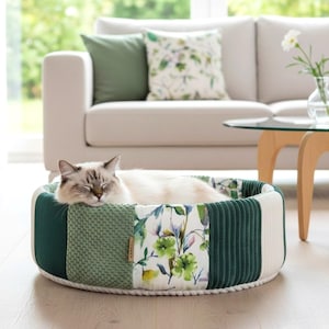 May include: A sleeping cat rests in a round pet bed. The bed features a patchwork design with green corduroy, textured green fabric, floral print, and white panels. The bed is on a wooden floor.